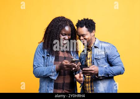 Les partenaires afro-américains envoient des messages en ligne sur leurs smartphones, regardant sur les médias sociaux devant la caméra. Jeunes gens engagés montrant des vidéos amusantes sur les téléphones. Banque D'Images