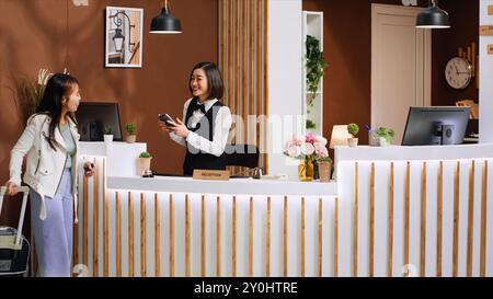 Voyageur asiatique payant pour la chambre d'hôtel lors du processus d'enregistrement, choisissant POS terminal paiement avec carte de crédit. Jeune femme avec des bagages faisant une transaction électronique pour l'hébergement. Banque D'Images