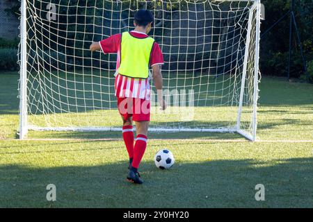Jouant au football, garçon en uniforme rouge donnant un coup de pied à la balle vers le but sur le terrain Banque D'Images