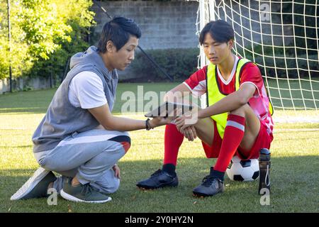 Entraîneur montrant la tablette à un adolescent joueur de football masculin assis sur le ballon pendant la séance d'entraînement Banque D'Images