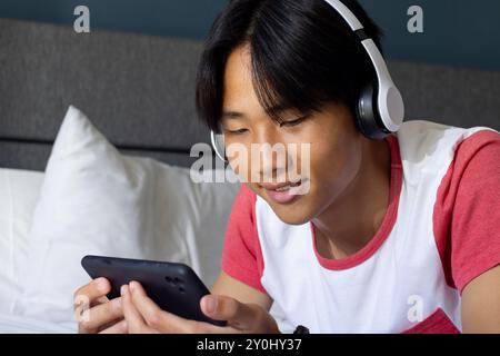 Écouter de la musique avec des écouteurs, jeune homme asiatique utilisant un smartphone sur le lit Banque D'Images