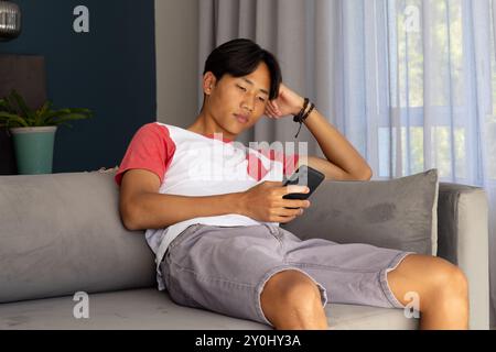 Détente sur le canapé, jeune homme asiatique naviguant smartphone, portant des vêtements décontractés Banque D'Images