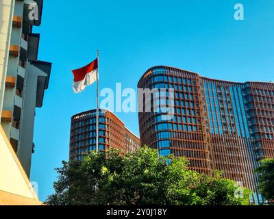 Drapeau indonésien agitant contre le ciel bleu clair avec immeuble de bureaux moderne à l'arrière-plan. Banque D'Images