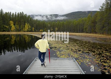 Une femme se tient debout sur un quai appuyé contre un parapluie rouge plié au lac Sinclair dans le nord de l'Idaho pendant l'automne Banque D'Images