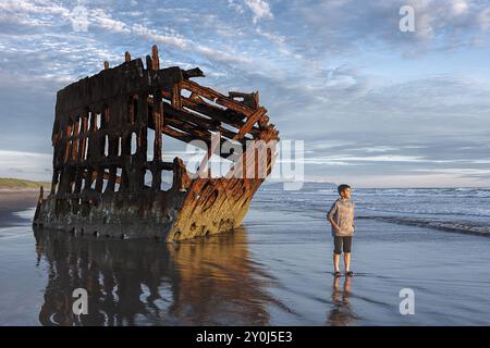 Boy se tient près du naufrage Peter Iredale dans la lumière dorée du coucher de soleil dans le nord-ouest de l'Oregon Banque D'Images