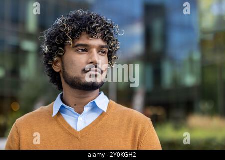 Jeune homme confiant avec des cheveux bouclés se tient à l'extérieur avec une expression réfléchie. Portant un pull orange décontracté et une chemise bleue, il respire le professionnalisme et la concentration. Banque D'Images