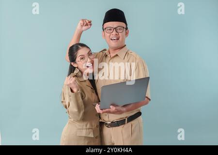 Deux employés du gouvernement indonésien excités portant un uniforme kaki tenant un ordinateur portable avec une expression heureuse et réussie. PNS et concept de fonctionnaire. Banque D'Images