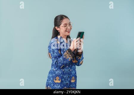Femme souriante employée du gouvernement indonésien portant un uniforme de batik bleu et des lunettes utilisant un smartphone pour naviguer sur Internet. PNS concept de fonctionnaire. Banque D'Images