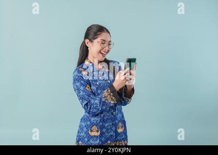 Femme souriante employée du gouvernement indonésien portant un uniforme de batik bleu et des lunettes utilisant un smartphone pour naviguer sur Internet. PNS concept de fonctionnaire. Banque D'Images