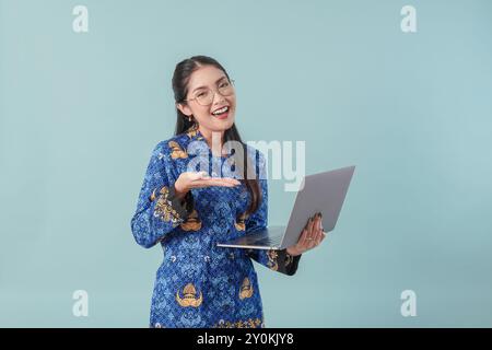 Femme joyeuse employée du gouvernement indonésien portant un uniforme de batik bleu et des lunettes pointant vers l'ordinateur portable qu'elle tient. PNS concept de fonctionnaire. Banque D'Images