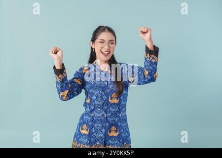 Femme travailleuse du gouvernement indonésien excitée portant un uniforme de batik bleu et des lunettes posant avec une expression heureuse et réussie. PNS concept de fonctionnaire Banque D'Images