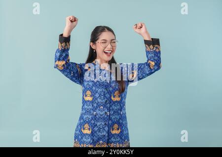 Femme travailleuse du gouvernement indonésien excitée portant un uniforme de batik bleu et des lunettes posant avec une expression heureuse et réussie. PNS concept de fonctionnaire Banque D'Images