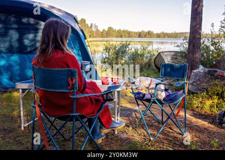 Femme, assise sur une chaise de camping devant une tente, appréciant une tasse de café et une vue imprenable en Norvège et en Suède le matin, camping sauvage. Femme Banque D'Images