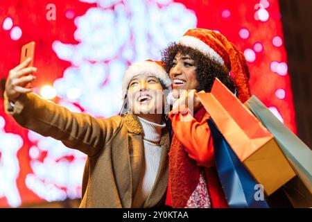 Deux amis heureux prenant selfie tout en tenant des sacs à provisions dans la nuit de noël Banque D'Images
