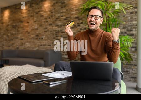 Homme d'affaires satisfait avec carte de crédit et ordinateur portable criant et célébrant la récompense à table dans le bureau Banque D'Images