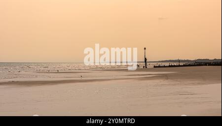 Une plage de sable, avec de l'eau couvrant le sable, a un brise-lames sur un côté. Les gens sont sur la plage et la mer scintille sous un ciel clair. Banque D'Images