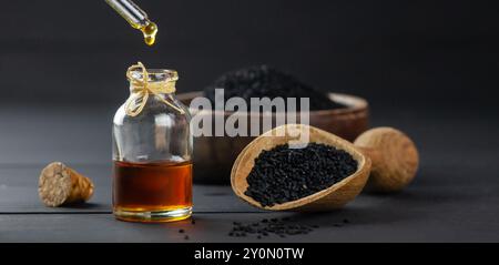 Bouteille en verre d'huile essentielle de graines de cumin noir, Nigella Sativa dans une cuillère sur fond de bois noir, goutte à goutte d'huile Banque D'Images