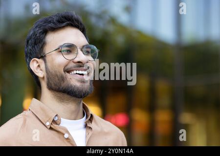 Portrait d'un jeune homme joyeux portant des lunettes, souriant chaleureusement dans un environnement urbain extérieur. Il montre confiance et bonheur, habillé en tenue décontractée. Banque D'Images