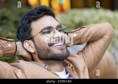 Le jeune homme souriant en tenue décontractée se détend à l'extérieur. Son expression joyeuse et sa posture confortable véhiculent des sentiments de bonheur et de loisirs. Image idéale pour les concepts de relaxation, positivité Banque D'Images