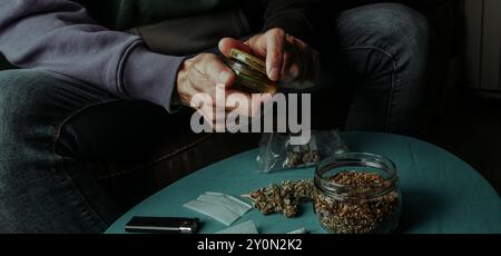 un homme déchiquette un bourgeon de cannabis avec un moulin, assis sur un canapé à côté d'une table avec du tabac à rouler et du papier, dans un format panoramique à utiliser comme bann web Banque D'Images