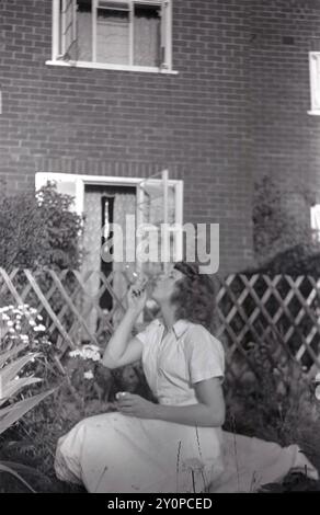 1960, historique, été et une jeune dame, assis dans un jardin arrière d'une maison de banlieue, souffler des bulles à travers un verre, Angleterre, Royaume-Uni. Banque D'Images