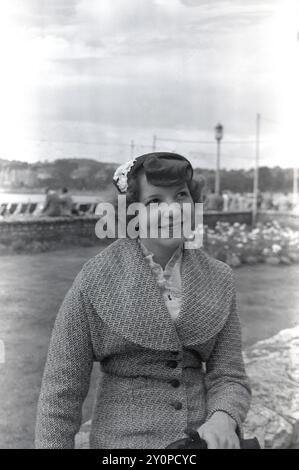 1955, historique, une jolie jeune femme, chemisier en dentelle, veste en laine à la mode avec des revers larges, et petit chapeau de fleur, assis dehors pour une photo, Angleterre, Royaume-Uni. Banque D'Images