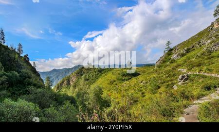 Panorama d'un paysage d'été ensoleillé dans les montagnes de l'Altaï. Un chemin d'enroulement étroit mène vers le haut. Une calotte de nuages blancs se trouve sur les sommets de la montagne Banque D'Images