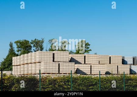 Grandes piles de palettes en bois disposées uniformément avec des pierres de béton dans une zone de stockage extérieure, avec des arbres et un ciel bleu clair en arrière-plan. Banque D'Images