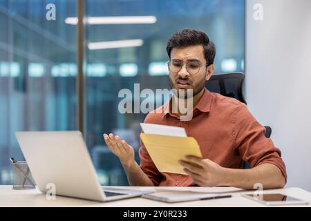 Jeune professionnel dans un environnement de bureau semblant frustré après avoir reçu de mauvaises nouvelles dans une enveloppe postale. Assis au bureau avec ordinateur portable, téléphone et documents. Banque D'Images