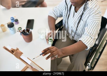 Personne assise dans un fauteuil roulant, tenant un pinceau et une toile de peinture sur un chevalet tout en se concentrant sur la création de multiples Diverses peintures et pinceaux sont dispersés dans l'espace de travail Banque D'Images
