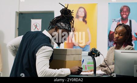 Gars afro-américain ramassant la commande au magasin de vêtements, signant des papiers de confirmation à la caisse enregistreuse. Femme employée préparant des boîtes avec des articles de mode et des accessoires demandés. Caméra A. Banque D'Images