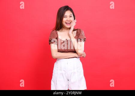 Expression de femme asiatique joyeuse avec ses bras croisés comme un signe, portant une blouse brune et un short sur un fond rouge. pour la mode, le style de vie et un Banque D'Images