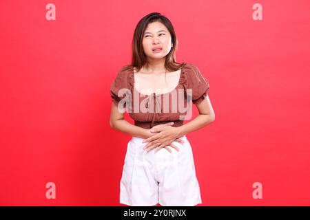 candide indonésie expression de la femme dans la douleur avec des problèmes d'estomac portant une blouse brune et un short sur un fond rouge. pour la santé, le style de vie et l'adv Banque D'Images