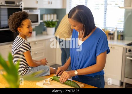 Mère coupant le concombre dans la cuisine tandis que la fille souriante et assise sur le comptoir Banque D'Images