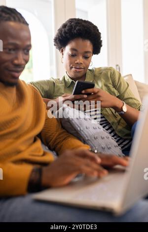 En utilisant smartphone et ordinateur portable, couple afro-américain se relaxant ensemble sur le canapé à la maison Banque D'Images