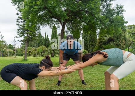 Deux femmes pratiquent l'étirement partenaire dans un parc verdoyant, supervisé par un entraîneur de fitness. La session favorise la flexibilité, le travail d'équipe et les avantages o Banque D'Images