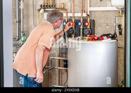 Technikraum einer Heizung, Teil einer neu installierten Wärmepumpenanlage, die eigentliche Wärmepumpe wurde draußen installiert, Glonn, Bayern, September 2024 Deutschland, Glonn, septembre 2024, Technikraum einer Heizung, Teil einer neu installierten Wärmepumpenanlage, die eigentliche Luft-Wärmepumpe steht draußen, hier im Keller befindet sich der Warmwasserspeicher für Heizung und Warmwasser und der Wärmetauscher verdeckt, links, Mann überprüft die Einstellungen, Rentner, die Anlage wurde erst vor zwei Tagen installiert, die installation noch nicht abgeschathen, Privlossen, Luftwärmepump,, Banque D'Images
