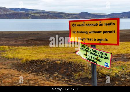 Un panneau sur un poteau dans un champ près d'un lac avec des montagnes derrière. Le panneau est jaune et rouge Banque D'Images