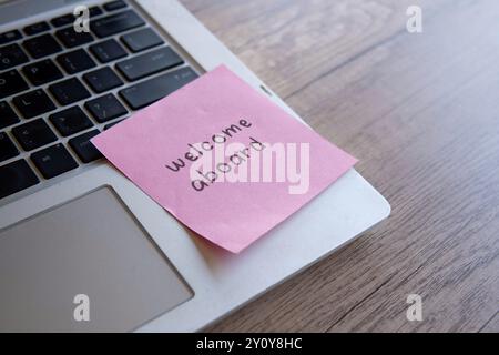 Un gros plan d’un clavier d’ordinateur portable avec une note autocollante rose attachée avec le texte « Bienvenue à bord », symbolisant un nouvel emploi, l’intégration en équipe Banque D'Images