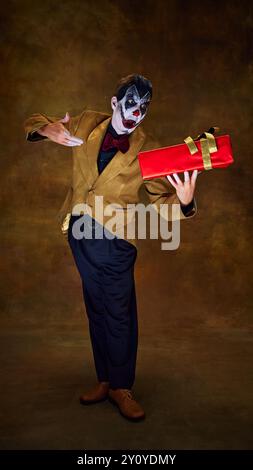 Tenant fièrement un cadeau rouge, le clown sourit malicieusement, comme s'il présentait quelque chose de dangereux sur fond sombre vintage. Banque D'Images