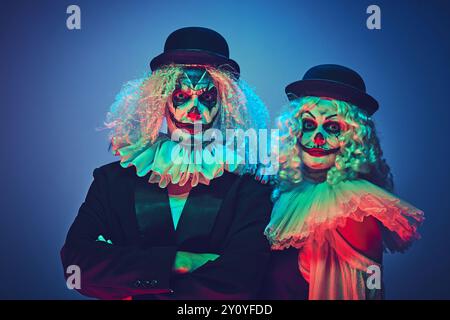 Portrait d'un couple de clown effrayant habillé des vêtements de style rétro posant regardant la caméra calmement dans la lumière néon sur fond dégradé. Banque D'Images