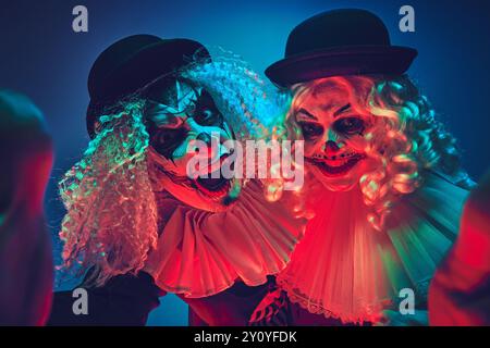 Portrait d'un couple de clown effrayant posant de manière ludique regardant la caméra dans la lumière néon sur fond dégradé. Banque D'Images