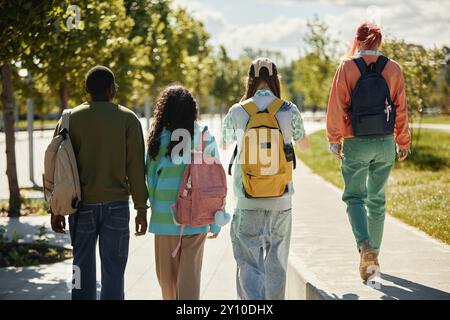 Vue arrière d'un groupe diversifié d'étudiants adolescents avec des sacs à dos habillés de vêtements décontractés lumineux marchant ensemble après les cours dans le parc de la ville le jour ensoleillé Banque D'Images