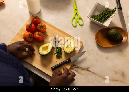 Préparer de l'avocat frais et des tomates sur une planche à découper, homme afro-américain hachant des herbes Banque D'Images