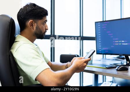 En utilisant un smartphone, homme indien assis à un bureau avec un écran d'ordinateur affichant le code Banque D'Images