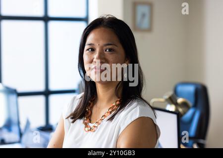 Femme souriante au bureau portant chemisier blanc et collier, debout avec confiance Banque D'Images