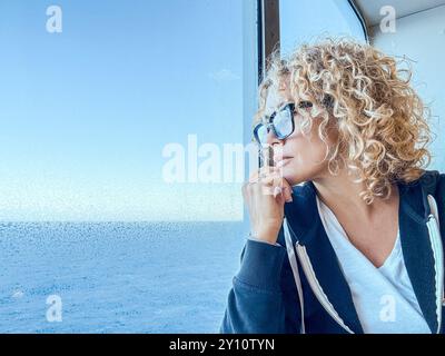 Belle femme avec des lunettes passant du temps libre à regarder la mer. Elle prend soin d'elle, se relaxant. Profitez de la vue, regardez par la fenêtre. La blonde d'âge moyen et la femme bouclée semblent réfléchies. Matins paisibles. Banque D'Images