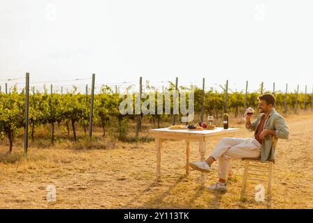 L'homme profite d'un moment de paix à une table rustique au milieu des rangées de vignes luxuriantes, en savourant du vin et des fruits frais tout en trempant dans la lumière dorée de l'après-midi Banque D'Images