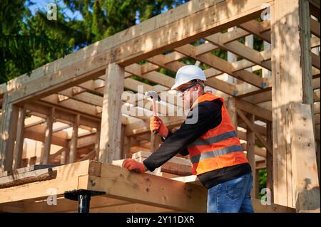 Charpentier construisant une maison à ossature en bois près de la forêt. Homme barbu avec des lunettes martelant des clous dans la structure, portant un casque de protection, gilet de construction. Concept de construction écologique moderne. Banque D'Images