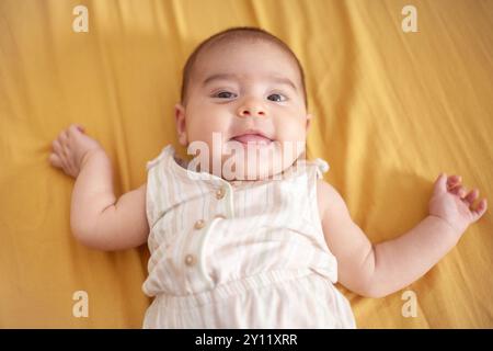 Un bébé est allongé sur un lit avec une couverture jaune. Le bébé sourit et sort la langue. Banque D'Images